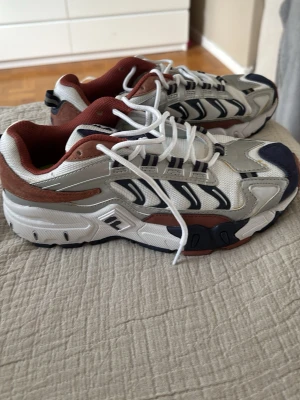 Vita, bruna och marinblå sneakers från Fila - Snygga sneakers från Fila i vitt, marinblått och brunt. Skorna har en chunky sula, mesh- och läderdetaljer samt vita skosnören. Fila-logga på plösen och coola kontrastpaneler på sidorna. Perfekta för dig som gillar sportig och trendig stil.