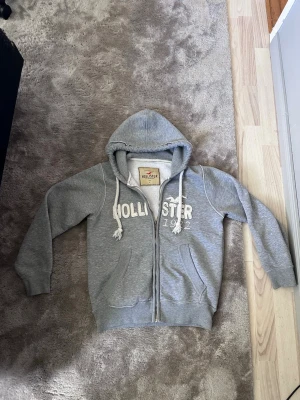 Vintage Hollister Zip Hoodie - Säljer en vintage grå hoodie från Hollister med dragkedja, stora fickor och vit broderad logga på bröstet. Storlek L men sitter verkligen som en M/ S