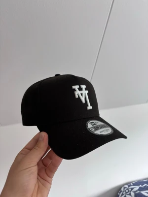 Svart New Era 9FORTY keps - Snygg svart keps från New Era, modell 9FORTY, med justerbar rem bak. Framtill finns en vit broderad logga med bokstäverna 'MT'. Kepsen har böjd skärm och klassisk passform. Tillverkad i slitstarkt tyg som passar perfekt till streetwear-stilen.