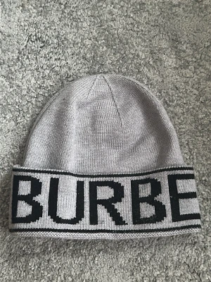 Grå Burberry mössa med logga - Snygg grå mössa från Burberry med bred uppvikt kant och stor svart logotyp runt hela mössan. Tillverkad i mjukt stickat material som sitter skönt på huvudet. Perfekt accessoar för att lyfta din outfit med en exklusiv touch.