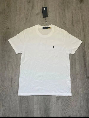 Vit t-shirt från Polo Ralph Lauren - Klassisk vit t-shirt från Polo Ralph Lauren med rund halsringning och den ikoniska lilla broderade loggan på bröstet. Tillverkad i mjuk bomull och har en normal passform. Perfekt basplagg för alla tillfällen.