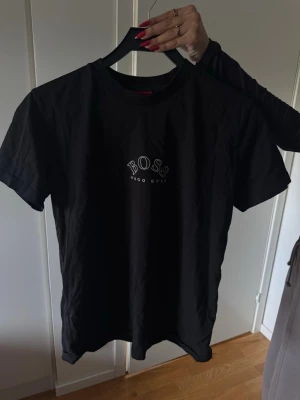 Svart Hugo Boss t-shirt med logga - Snygg svart t-shirt från Hugo Boss med stor vit logga både fram och bak. Klassisk rund hals och korta ärmar. Tillverkad i mjuk bomull som känns skön mot huden. Perfekt för dig som gillar stilrena och trendiga plagg.