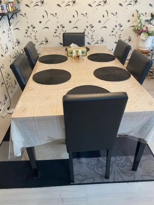 Matbord med 6 svarta stolar - Säljer ett stort rektangulärt matbord med ljus duk och plastskydd, samt 6 svarta stolar med skinnimitation. Bordet har plats för många och passar perfekt för middagar eller spelkvällar. 