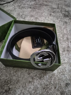 Svart skärp med silverdetalj från Gucci - Snyggt svart skärp från Gucci med ikoniskt dubbelt G-spänne i silver. Skärpet är gjort i slätt skinn och levereras med originalförpackning, dustbag och certifikat. Perfekt accessoar för att lyfta din outfit.