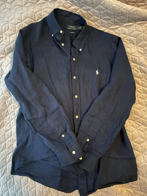 Marinblå linneskjorta Polo Ralph Lauren - Snygg marinblå skjorta från Polo Ralph Lauren i linne med klassisk button-down krage och vit broderad logga på bröstet. Skjortan har långa ärmar och vita knappar framtill. Perfekt för en stilren och avslappnad look.