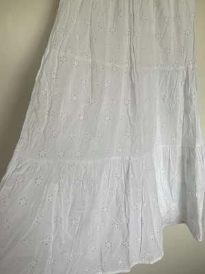 Vit långkjol med broderade blommor Gina tricot - Säljer en vit långkjol med broderade blommor och volangdetaljer. Kjolen har en luftig och flowy passform, perfekt för varma dagar. Materialet känns lätt och andas, vilket gör den skön att bära under sommaren. Jag rekommenderar passar perfekt till vuxen XS/S👌