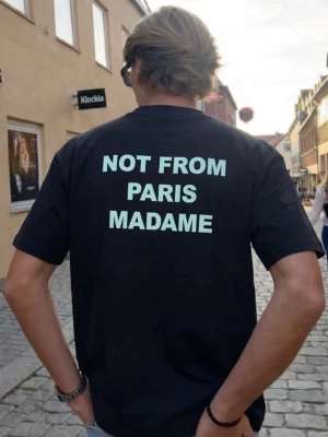 Svart t-shirt NOT FROM PARIS MADAME - Cool svart t-shirt med trycket 'NOT FROM PARIS MADAME' i vitt på ryggen. Klassisk passform och rund hals. Perfekt för dig som gillar streetwear och vill sticka ut med ett statement-plagg.