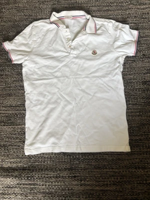 Vit Moncler piké med röd detalj - Klassisk vit pikétröja från Moncler med korta ärmar och knappar framtill. Kragen har snygga röda och blå ränder och Moncler-loggan är broderad på bröstet. Tillverkad i bomull för en skön och luftig känsla.