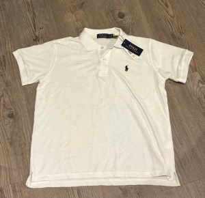 Vit pikétröja från Polo Ralph Lauren - Klassisk vit pikétröja från Polo Ralph Lauren med korta ärmar och krage. Tröjan har två knappar vid halsen och den ikoniska broderade loggan på bröstet. Perfekt till jeans eller shorts för en clean och stilren look. Är storlek L men passar mer M