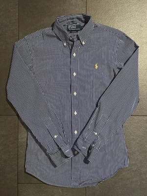 Randig blå skjorta från Polo Ralph Lauren - Säljer en klassisk blå- och vitrandig skjorta från Polo Ralph Lauren i slim fit. Skjortan har långa ärmar, knappar framtill och den ikoniska gula logotypen broderad på bröstet. Perfekt för dig som gillar stilrena och tidlösa plagg.
