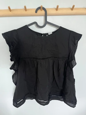 Svart blus med volangdetaljer - Svart blus i luftig bomull med volangdetaljer längs ärmarna och dekorativa broderade hål vid midja och fåll. Rund halsringning och korta ärmar ger en avslappnad men stilren look. Perfekt att matcha med jeans eller kjol. Aldrig använd. Pris kan diskuteras!🥰storlek M men skulle säga att den passar S också