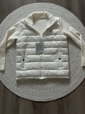 Moncler cardigan - Snygg vit dunväst från Moncler med stickade ärmar och hög krage. Västen har två framfickor med knappstängning och Moncler-logga på bröstet. Materialet är en kombination av dun och stickad bomull vilket ger en modern och sportig look.