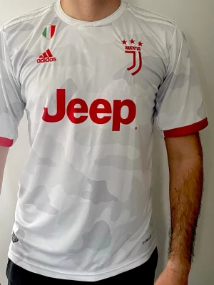Juventus bortatröja Ronaldo 7 Adidas - Juventus bortatröja i vitt med ljusgrått camomönster, röda detaljer och tryck. Adidas-logga, klubbmärke och Jeep sponsor på framsidan. RONALDO och nummer 7 på ryggen. Tillverkad i lätt och ventilerande material, perfekt för fotboll eller träning. Säsong 2019/202. Storlek är L.