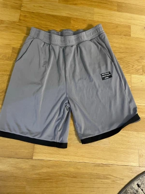 Grå shorts med svart kant - Snygga grå shorts med svart kant längst ner och resår i midjan. Har sidofickor och en liten svart patch med text på vänster ben. Perfekta för en avslappnad stil.