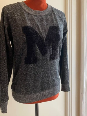 Mörkgrå sweatshirt med M-applikation XS - Tröja med rund halsringning och långa ärmar. Tröjan är gråmelerad med ett stort "M" i frotté framtill. Längd: 59 cm Bystvidd: 48 cm 80% polyester 20% bomull  Inga bilder på mig! Katter finns i hemmet!