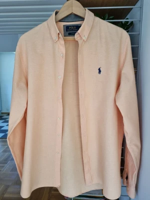 Persikofärgad skjorta från Polo Ralph Lauren - Obs står XL! Dock liten stl mer som stl L/M Snygg persikofärgad skjorta från Polo Ralph Lauren med klassisk button-down krage och broderad marinblå logga på bröstet. Skjortan har långa ärmar och är tillverkad i ett mjukt bomullstyg. Perfekt för en stilren look.