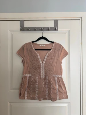 Odd molly Rosa blus med broderi och kort ärm - Söt gammelrosa blus med vita broderade detaljer framtill och längs midjan. Blusen har v-ringning, korta ärmar och ett lätt, luftigt tyg som ger en avslappnad känsla. Perfekt till jeans eller kjol för en trendig look. Storlek S