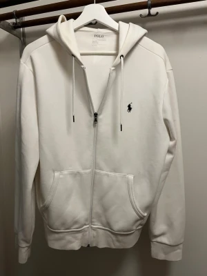 Vit hoodie från Polo Ralph Lauren - Snygg vit hoodie från Polo Ralph Lauren med dragkedja, huva med dragsko och två stora fickor framtill. Klassisk logga broderad på bröstet. Perfekt för en clean och stilren look.