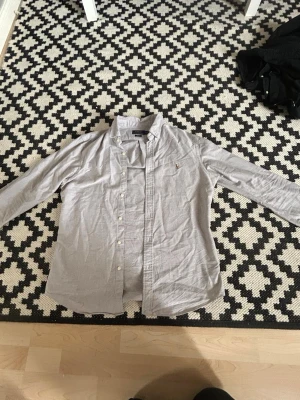 Ljusgrå skjorta från Polo Ralph Lauren - Klassisk ljusgrå skjorta från Polo Ralph Lauren med knappslå och broderad logga på bröstet. Skjortan har lång ärm, button-down krage och är tillverkad i mjuk bomull. Perfekt till jeans eller chinos för en clean look.