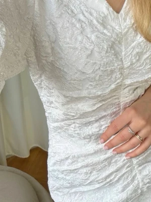 Vit kort omlottklänning BYIC - Supersnygg vit kort klänning från BYIC med puffärmar och omlottdesign. Klänningen har en strukturerad yta och v-ringning, vilket ger en trendig och elegant look. Perfekt för dig som vill sticka ut med stilrena detaljer och modern passform. Den är använd en gång för 2 år sedan. 