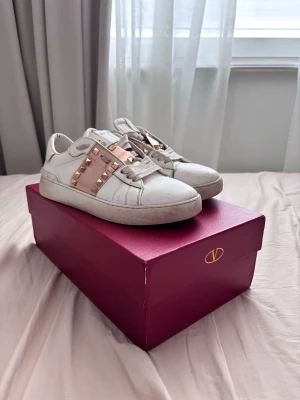 Valentino Garavani vita sneakers - Snygga vita sneakers från Valentino Garavani med breda metallicrosa band och guldfärgade nitar över vristen. Skorna är låga och har klassisk snörning samt detaljer i läder. Perfekta för dig som vill sticka ut med exklusiva detaljer.