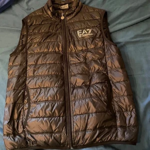 Svart dunväst från Emporio Armani - Snygg svart dunväst från Emporio Armani med hög krage och dragkedja framtill. Västen har EA7-logga på bröstet och två sidofickor med dragkedja. Lättviktigt material och quiltad design gör den både trendig och bekväm.