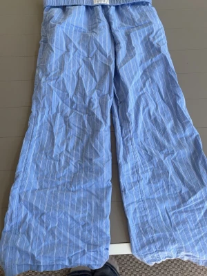 Blåa pyjamasbyxor med vita ränder - Mysiga blåa pyjamasbyxor med tunna vita vertikala ränder. Byxorna har en avslappnad passform och resår i midjan för extra komfort. Perfekta för chillkvällar hemma eller när du bara vill ta det lugnt.