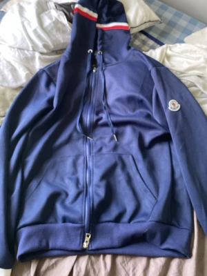 Moncler hoodie - (HELT NY) Snygg marinblå hoodie från Moncler med dragkedja framtill och fickor på sidorna. Huva med dragsko och rödvit randdetalj upptill. Moncler-logga på vänster ärm. Tillverkad i mjukt bomullsmaterial, perfekt för en avslappnad stil.