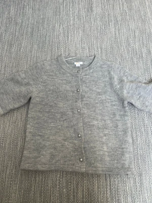 Grå kofta från Gina Tricot - Mysig grå kofta från Gina Tricot med rund halsringning och pärlknappar framtill. Koftan är stickad i mjukt material och har korta ärmar. Perfekt att slänga på sig över en t-shirt eller linne.