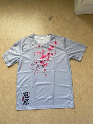 Ljusgrå t-shirt med körsbärsblommor - Snygg ljusgrå t-shirt med korta ärmar och ett stort tryck av rosa och svarta körsbärsblommor på framsidan. T-shirten har rund halsringning och japansk text nertill. Tillverkad i ett lätt och mjukt syntetmaterial som känns skönt mot huden.