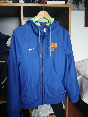 Blå FC Barcelona Nike jacka med huva - Snygg blå FC Barcelona jacka från Nike med klubbmärke på bröstet och limegröna detaljer. Jackan har huva med dragsko, hel dragkedja och två fickor med dragkedja. Perfekt för träning eller när du vill representera ditt lag.