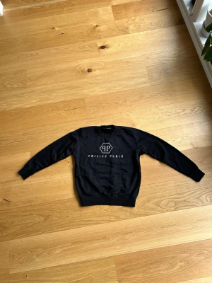 Svart sweatshirt från Philipp Plein. - Svart sweatshirt från Philipp Plein med vit logga och text på bröstet. Tröjan har rund hals, långa ärmar och ribbade muddar vid ärmslut och nederkant. Materialet är mjukt och bekvämt, Det står S men det passar xs, själv har jag xs.