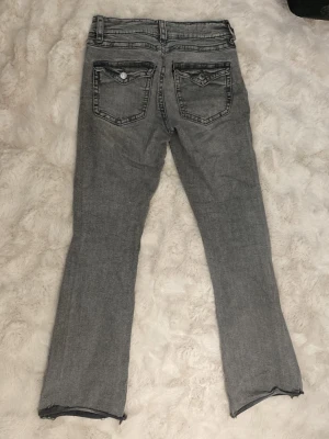Grå flared jeans med rå kant - Säljer ett par grå flared jeans , avklippta benslut. Jeansen har fem fickor, dekorativa sömmar och knäppning fram. Snygg tvättad look och detaljerade bakfickor med lock och knapp.