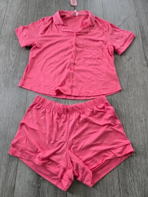 Rosa pyjamasset med kortärmad skjorta S - Säljer ett rosa pyjamasset med kortärmad skjorta och matchande shorts. Skjortan har knappar framtill, krage och en bröstficka. Shortsen har resår i midjan för extra komfort. Materialet är mjukt och lätt, perfekt för mysiga kvällar.
