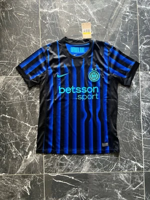 Inter Milan matchtröja Nike blå/svart - Snygg Inter Milan matchtröja från Nike med klassiska blå och svarta vertikala ränder. Tröjan har korta ärmar, rund hals och sponsorlogga 'betsson sport' på bröstet. Materialet är lätt och andas, perfekt för fotboll eller träning.