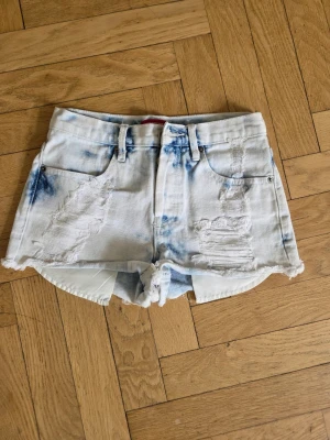 Ljusa slitna jeansshorts  - Säljer ett par ljusa jeansshorts stl 26 med slitna detaljer och blå tie-dye-effekt. Shortsen har råa kanter, fem fickor och bälteshällor. Perfekta för sommaren och ger en avslappnad vibe till din outfit.MÅTT FRÅN GRENEN OCH UPP CA 26 CM , MIDJEMÅTT RAKT ÖVER CA 33 CM , KNAPPAR I GYLFEN 