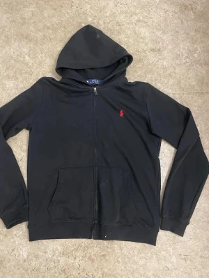 Svart hoodie från Polo Ralph Lauren - Klassisk svart hoodie med dragkedja från Polo Ralph Lauren. Har huva, känguruficka och den ikoniska röda logotypen broderad på bröstet. Tillverkad i mjukt bomullsmaterial och har ribbade muddar vid ärmslut och nederkant.