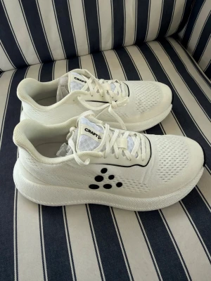 Vita sneakers från Craft - Stilrena vita sneakers från Craft med svart logotyp på sidan och svarta detaljer runt snörningen. Ovandelen är i ventilerande meshmaterial och sulan är tjock och ger bra dämpning. Perfekta för en sportig och modern look.