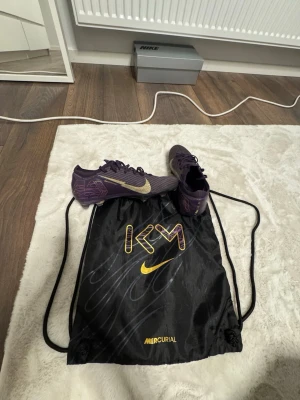 Nike Mercurial KM lila fotbollsskor - Säljer ett par Nike Mercurial KM fotbollsskor i lila med guldfärgade detaljer och swoosh. Skorna har låg profil, snörning och är tillverkade i syntetmaterial för bästa bollkontroll. Medföljer svart gympåse med KM-logga och Mercurial-tryck. Perfekta för snabba spelare. Ny pris 3500 lite slitet där på toppen av snöret men annars i bra skick.