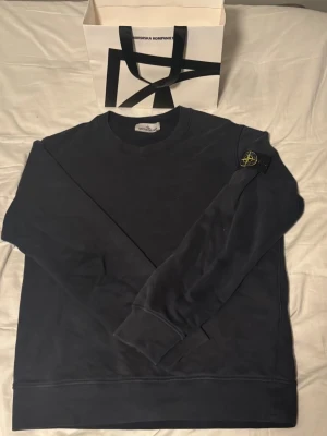Mörkblå sweatshirt från Stone Island - Mörkblå sweatshirt från Stone Island med klassisk rund halsringning och ribbade muddar. Tröjan har den ikoniska Stone Island-loggan på vänster ärm. Tillverkad i mjukt bomullsmaterial och har en avslappnad passform.