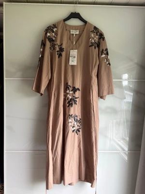 Beige långklänning med paljettdekoration Munthe - Säljer en beige långklänning från Munthe med v-ringning och trekvartsärm. Klänningen har vacker paljettdekoration i svart och vitt på axlar, ärmar och framtill. Tillverkad i bomull och har en avslappnad passform. Passar alla från XS-L beroende på hur oversize man vill ha den. Ca 118cm lång. Ord. pris 3900kr.