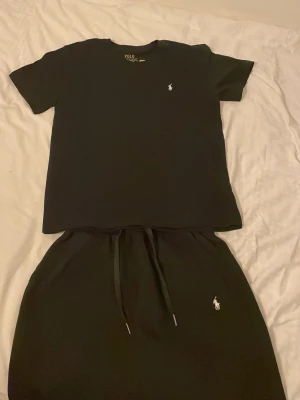 Svart set från Polo Ralph Lauren - Snyggt svart set från Polo Ralph Lauren med klassisk t-shirt och matchande shorts. Båda plaggen har den ikoniska vita polospelaren broderad på bröstet och benet. Mjukt och bekvämt material, perfekt för en avslappnad stil.