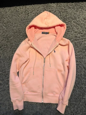 Ljusrosa hoodie från Polo Ralph Lauren - Snygg ljusrosa hoodie med dragkedja från Polo Ralph Lauren. Klassisk design med huva, dragsnören och två fickor framtill. Liten broderad logga i blått på bröstet. Mjuk bomullsblandning och ribbade muddar vid ärmslut och nederkant.