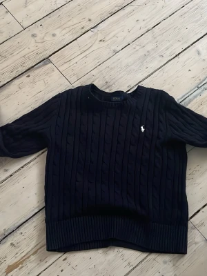  Polo Ralph Lauren - Snygg mörkblå kabelstickad tröja från Polo Ralph Lauren med klassisk rund halsringning och vit broderad logga på bröstet. Tröjan har ribbade muddar och nederkant för en schysst passform. Perfekt för dig som gillar tidlös och stilren design.