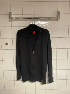 Svart ziptröja från Esprit - Svart stickad ziptröja från Esprit med ribbade ärmar och hög krage. Tröjan har hel dragkedja framtill och breda muddar vid ärmslut och nederkant. Perfekt för lager-på-lager och snygg att matcha med jeans.