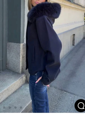 Mörkblå fluff kappa   med pälskrage - Snygg mörkblå kappa  med stor, fluffig pälskrage och raka ärmar. Jackan har en kortare modell och två fickor framtill. Perfekt för dig som vill ha en stilren och trendig look.