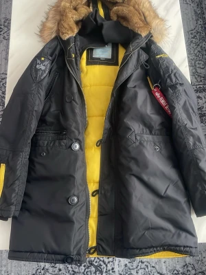 Svart parkas Alpha Industries M - Säljer en svart parkas från Alpha Industries med gul vadderad insida och avtagbar fuskpäls på huvan. Jackan har flera fickor, patch på ärmen och klassisk röd tag. Perfekt för kalla dagar och riktigt snygg streetstil. Vid frågor eller intresse skicka gärna ett meddelande så hörs vi!.