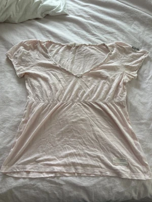 Odd Molly topp - Supersöt ljusrosa blus från Odd Molly med v-ringad hals och korta ärmar. Blusen har en feminin passform med resår i midjan och små spetsdetaljer längs halsringningen. Perfekt för en söt och avslappnad look.