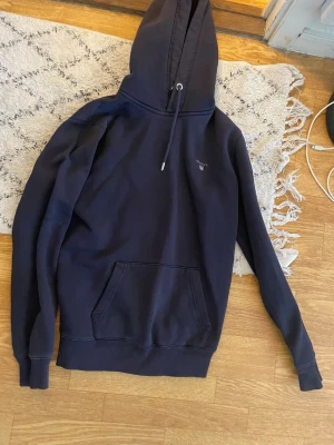 Mörkblå hoodie från GANT - Säljer en mörkblå hoodie från GANT med klassisk huva och dragsnören. Tröjan har en stor magficka och diskret GANT-logga på bröstet. Perfekt för dig som gillar stilrena och bekväma plagg. Materialet är mjukt och känns riktigt skönt mot huden.