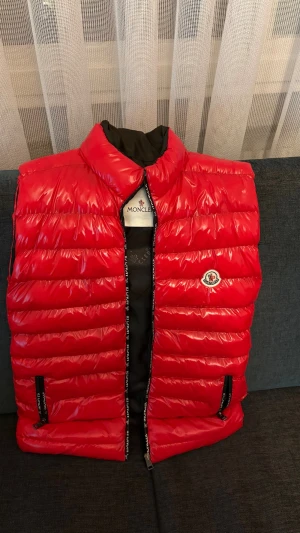 Röd dunväst från Moncler - Snygg röd dunväst från Moncler med glansig finish och klassisk logga på bröstet. Västen har hög krage, två dragkedjeförsedda fickor och quiltad design. Perfekt för lager-på-lager och ger en sportig vibe till din outfit.